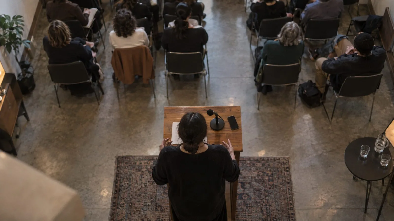 Anonymous speaker addressing an audience beside a smartphone and microphone / TokenPost.ai